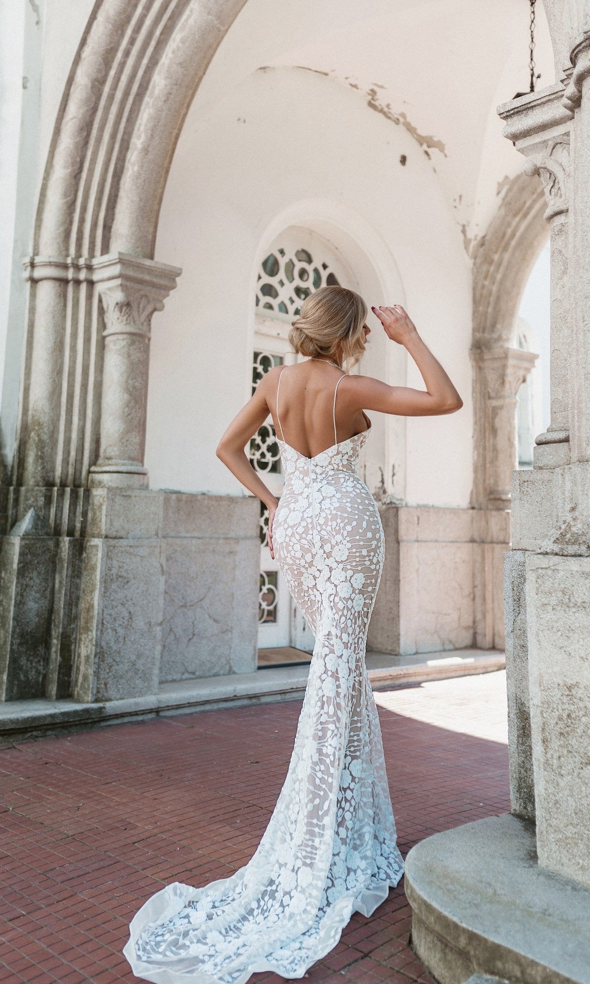 A woman in the Tina Holly BB008 long white lace formal dress stands outdoors under a stone arch, facing away and adjusting her hair.